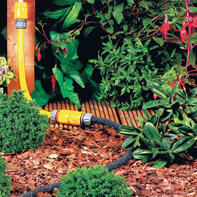 A yellow garden hose attached to a black soaker hose waters plants and shrubs in a garden with mulch and green foliage, surrounded by various leafy and flowering plants.
