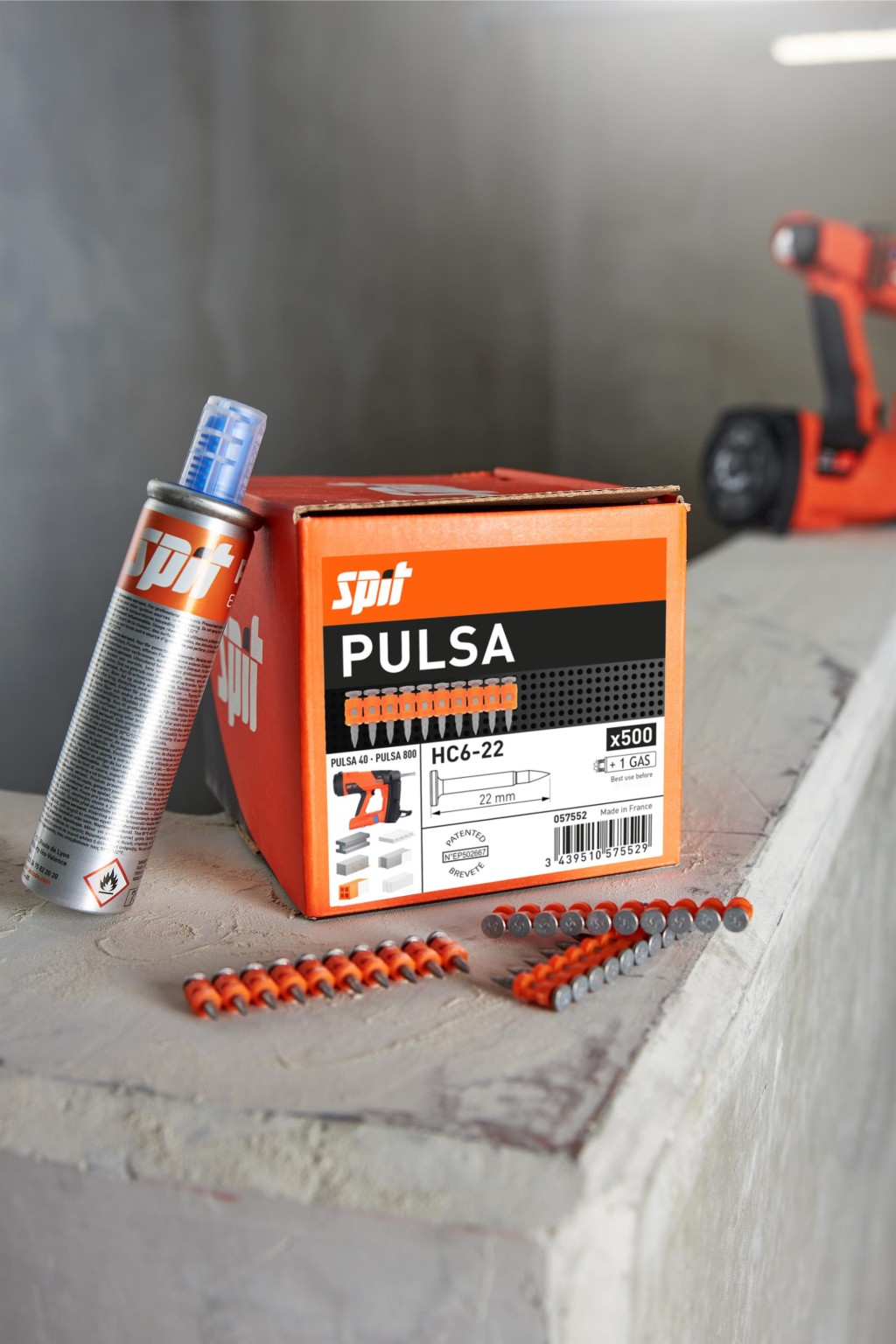 A box labeled Spit Pulsa with nails, a fuel cell, and nail strips on a concrete surface. An orange tool is blurred in the background. The packaging displays product specifications and branding.
