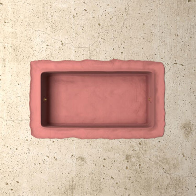 A rectangular, pink-colored sink with uneven, sculpted edges is set into a textured, light beige concrete surface. The sink has a matte finish and no visible fixtures.