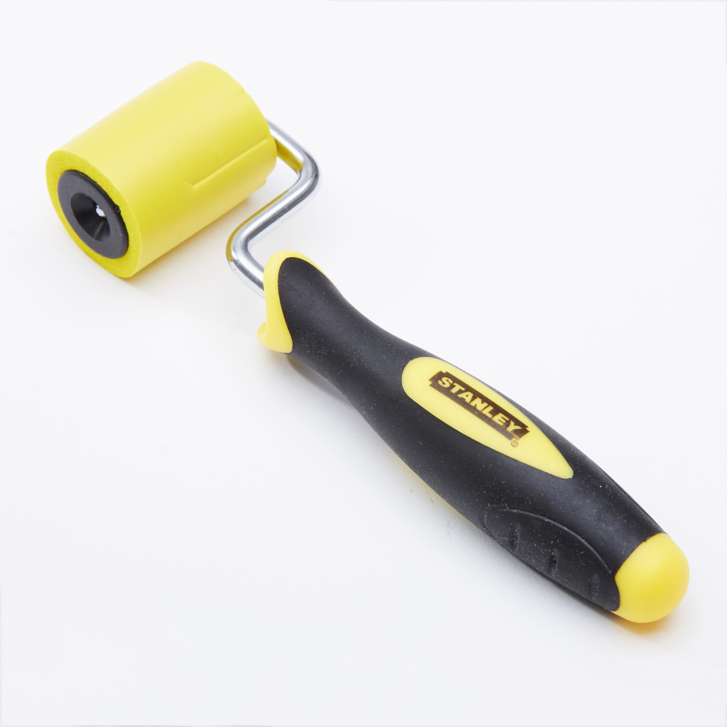 A yellow and black Stanley brand hand roller with a rubber handle and a cylindrical foam roller, placed on a white background.