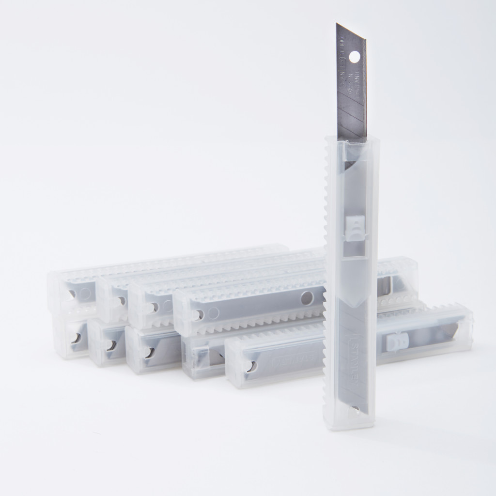 Several transparent plastic cases containing replacement utility knife blades are arranged in a row, with one case standing upright and displaying a blade partially extended from the top.