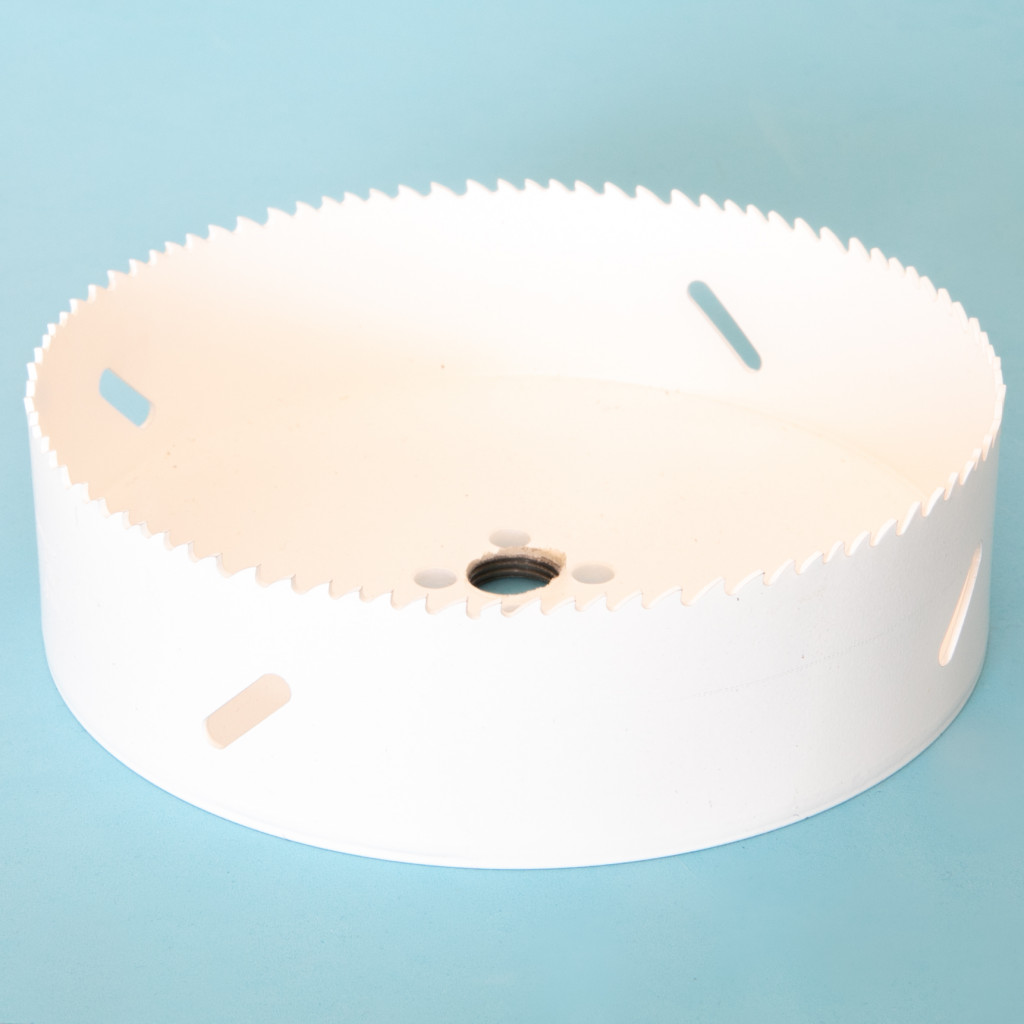 A white circular hole saw drill bit with sharp teeth along the edge and several elongated holes in the side, placed on a light blue background.