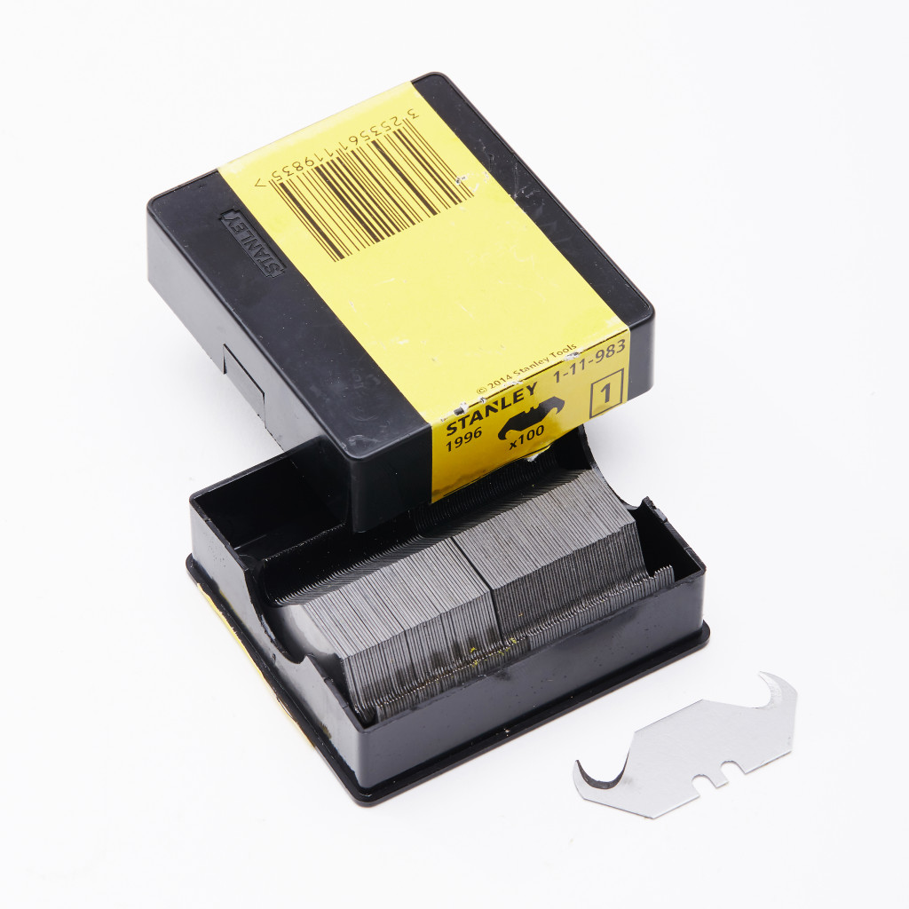 A black and yellow Stanley box holds a stack of utility knife blades, with one blade placed in front of the open box. A barcode is visible on the box lid.