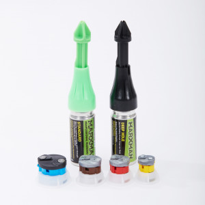 Two Markman marking tools, one green (standard) and one black (deep hole), stand upright. In front of them are four circular colored components: blue, brown, yellow, and red, resting on a white surface.