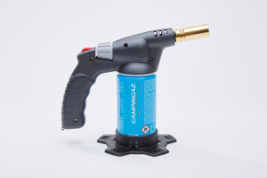 A portable butane gas torch with a black handle, blue gas canister labeled CAMPINGAZ, and a metallic nozzle, standing upright on a black plastic base against a white background.