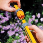 Two hands connect yellow garden hoses with gray and yellow plastic fittings, with purple flowers and green foliage blurred in the background.