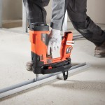 A person wearing gray work gloves and pants uses an orange cordless nail gun to fasten a metal strip to a concrete floor at a construction site.,