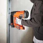A person wearing a dark jacket and white gloves uses an orange cordless nail gun to secure a metal stud to a wall. The scene appears to be part of a construction or renovation project.,