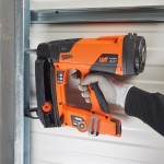 A person wearing a gray glove uses an orange and black Spit Pulsa 40P nail gun to fasten a metal frame to a wall with horizontal paneling.,