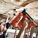A person wearing safety glasses uses an orange Paslode nail gun to secure wooden beams to a ceiling with insulation in a building under construction.,