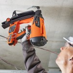Person wearing safety glasses and gloves using an orange and black nail gun to fasten materials to a concrete ceiling. The person’s face is partially visible and the tool is held overhead.,