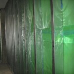 A row of metal wall studs covered with translucent green plastic sheeting, part of a construction or renovation project. The materials are arranged vertically along a bare wall.