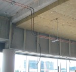 Exposed electrical conduit and wiring suspended from a concrete ceiling in a building under construction, with large windows and glass panels in the background.