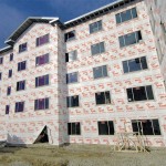 A multi-story building under construction, covered in Tyvek house wrap with exposed window openings, sits on a dirt lot under a clear blue sky.