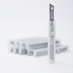 Several transparent plastic cases containing replacement utility knife blades are arranged in a row, with one case standing upright and displaying a blade partially extended from the top.