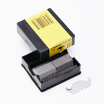 A black and yellow Stanley box holds a stack of utility knife blades, with one blade placed in front of the open box. A barcode is visible on the box lid.