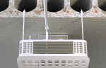 A close-up of a white ceiling-mounted air ventilation or HVAC unit, attached to a ceiling with visible round holes and exposed terrazzo-like material. Two metal support rods hold the unit in place.