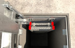 View from above of a metal ladder descending into a square concrete shaft. The top ladder rungs are highlighted with red markings, and the shaft walls have metal brackets attached.