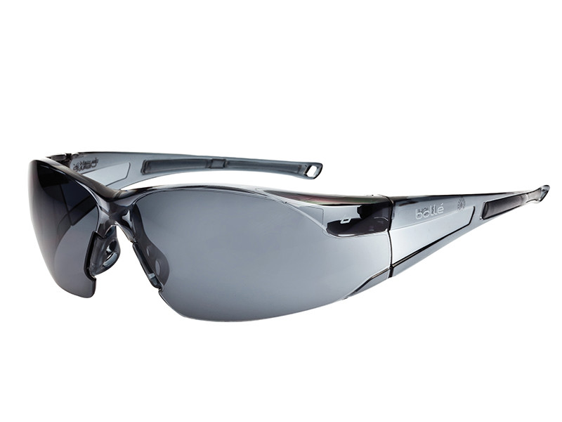A pair of sleek, wraparound safety glasses with dark-tinted lenses and a translucent, grayish-black frame. The brand name Bollé is visible on the temple.