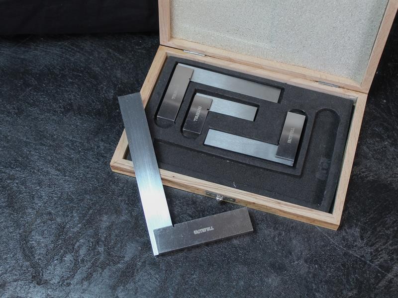 A wooden box with foam lining contains a set of metal engineer’s squares; one square is taken out and placed on a dark surface in front of the box.,