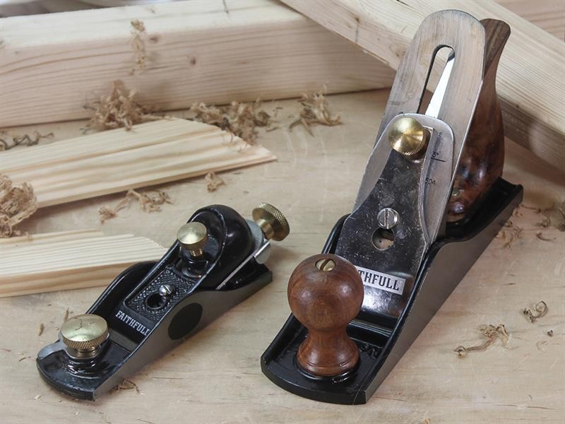 Two metal hand planes with wooden handles are placed on a workbench, surrounded by wood shavings and pieces of unfinished lumber. The tools are used for smoothing and shaping wood surfaces.