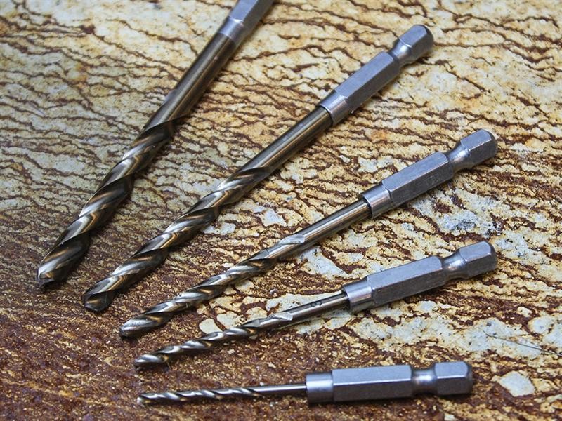 Five metal drill bits of different sizes are arranged on a textured, brownish surface resembling stone or wood grain. The drill bits have hexagonal shanks and sit parallel to each other.