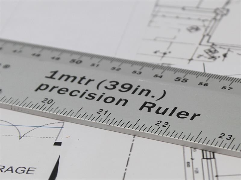 A close-up of a silver precision ruler labeled 1mtr (39in.) precision Ruler placed on top of technical drawings with measurement markings and diagrams visible underneath.,