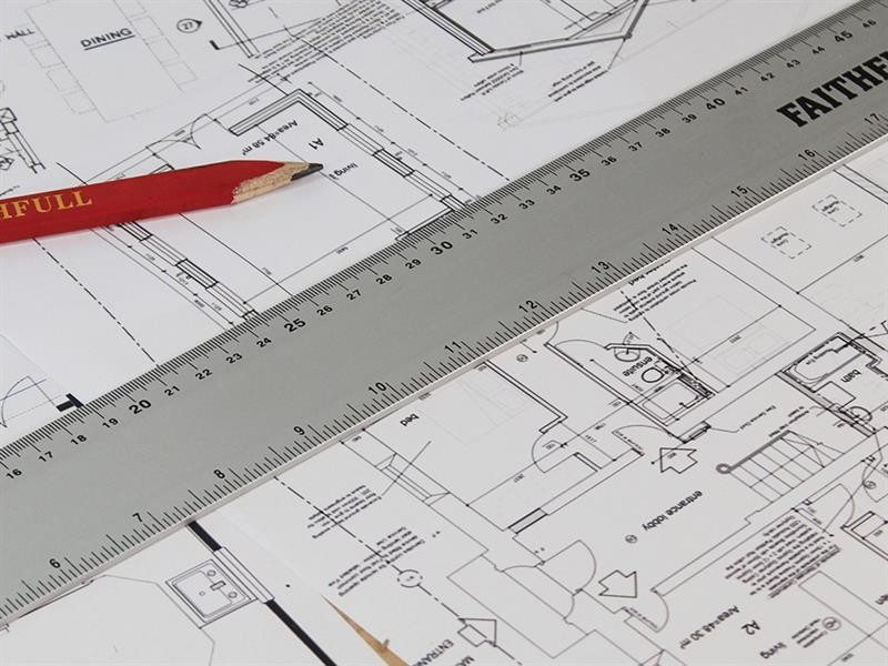 A close-up of architectural blueprints with a metal ruler and a red pencil placed on top, showing detailed building plans, measurements, and room layouts.
