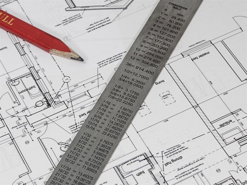 A metal ruler with measurement conversions and a red pencil rest on detailed architectural blueprints, showing floor plans and room layouts for a building.