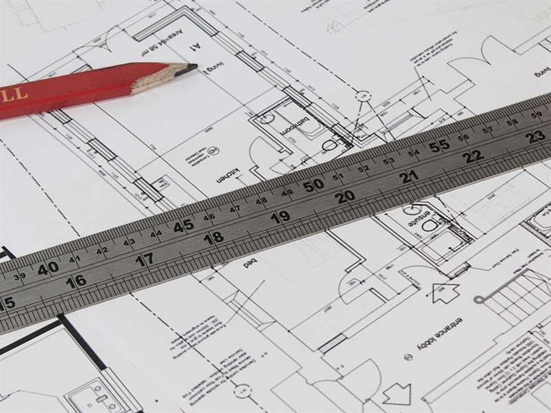 A close-up of architectural blueprints with a red pencil and a metal ruler placed on top, showing measurements and technical drawings of building plans.