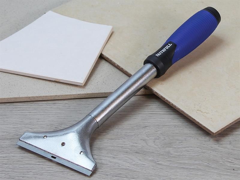 A heavy-duty scraper with a blue and black handle rests on a wooden surface next to beige ceramic tiles and a white tile.,