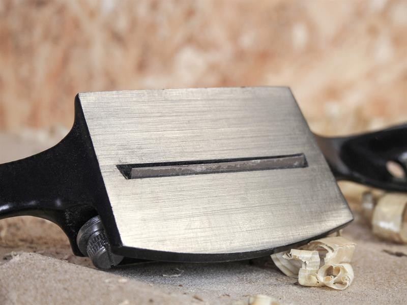 A close-up of a metal hand planer with wood shavings nearby, emphasizing its sharp blade and smooth surface. The background is blurred, highlighting the tools details.