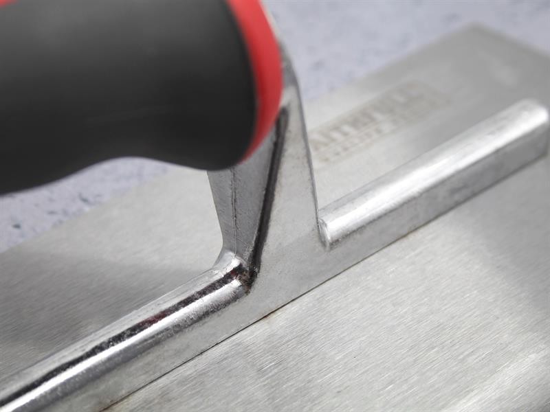 Close-up of a metal trowel with a black and red handle, resting on a smooth gray surface. The focus is on the handle and the joint where it meets the flat metal base.