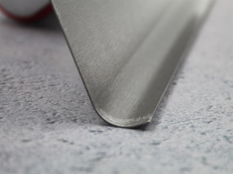 Close-up of the edge of a metal spatula resting on a light gray textured surface. The focus is on the smooth, beveled metal edge.