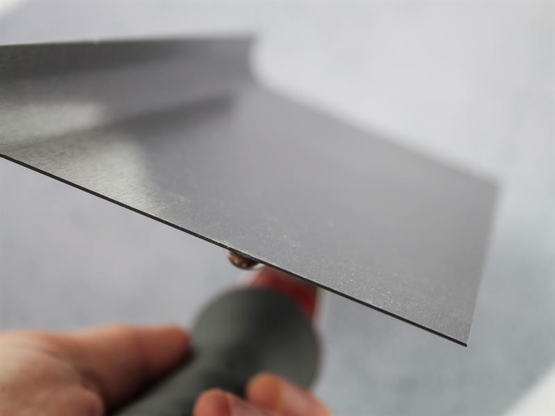A close-up of a person holding a metal drywall taping knife with a red and black handle, showing the sharp edge against a blurred, light background.