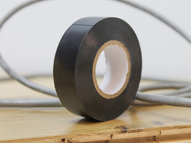 A roll of black electrical tape sits upright on a wooden surface with a gray electrical cable in the background.,