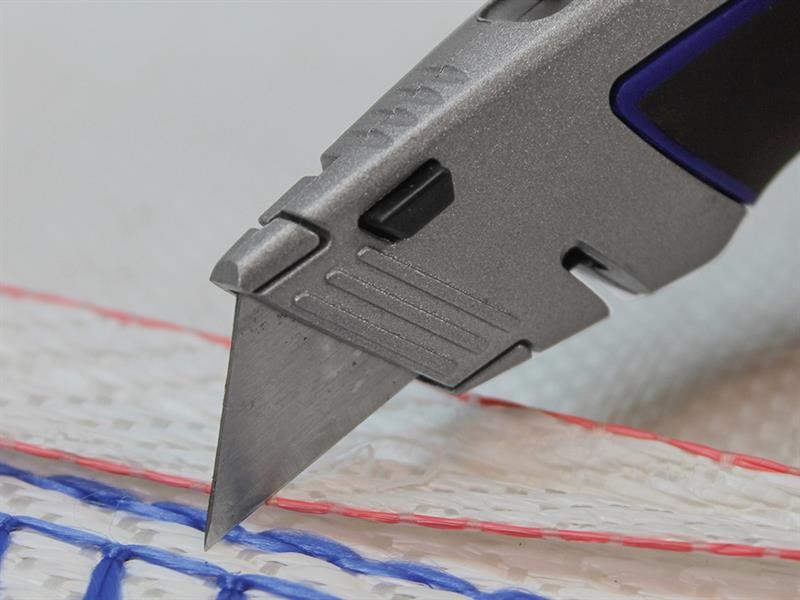 A close-up of a utility knife cutting through a woven white material with red and blue stitching.,