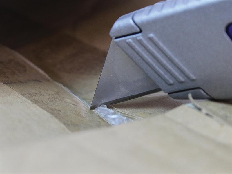 A close-up of a utility knife cutting through brown packing tape on a cardboard box.,