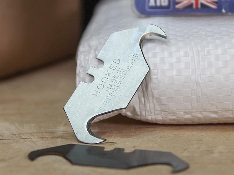A hooked utility blade with two curved, pointed ends rests against a woven white package. “HOOKED MADE IN SHEFFIELD ENGLAND” is engraved on the blade. Another similar blade lies flat on the surface nearby.