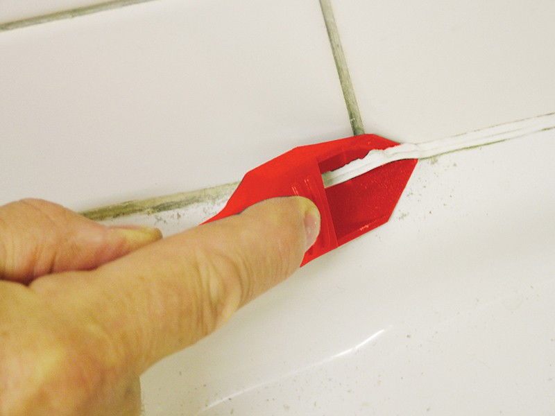 A hand uses a red plastic tool to apply or smooth white caulk along the seam between white bathroom tiles and a bathtub.,