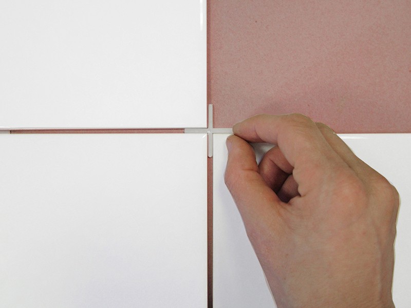 A hand places a tile spacer between four white tiles arranged on a pink surface, aligning them evenly for installation.