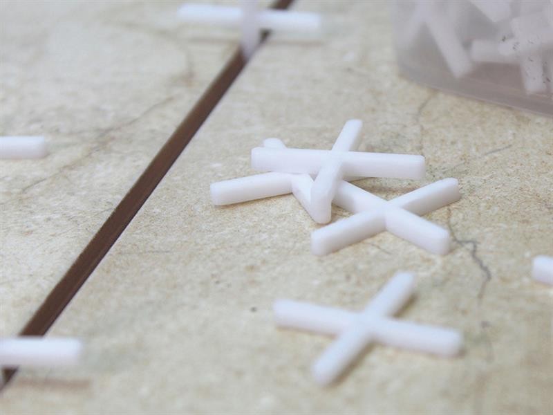 White plastic tile spacers are scattered on a beige tiled surface, with one spacer partially placed in the gap between two tiles. A plastic container with more spacers is visible in the background.