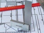A metal engineers square marked FAITHFULL stands upright on architectural blueprints, with a red pencil and a steel ruler in the background.