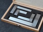 A wooden case with a foam insert holds five precision metal rulers of varying sizes, arranged neatly inside. The lid of the case is open, revealing the tools on a dark surface.