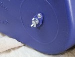 Close-up of a small metal bolt with a wing nut fastened onto a blue plastic surface, with part of a beige fabric visible underneath.