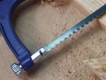 A close-up of a hacksaw with a blue handle cutting into a piece of wood. Wood shavings and sawdust are scattered on the wooden surface.,