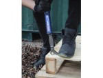 A person wearing black gloves and boots uses a blue crowbar to pry apart wooden pallets outdoors, next to a green metal structure and dried leaves on the ground.