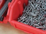 A close-up of a red plastic container filled with metal nails, sitting on a light wooden surface. Another identical, partially visible container is next to it.