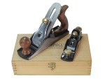 Two metal hand planes with wooden handles are placed on top of a closed wooden box. One plane is larger than the other, and both have brass adjustment knobs. The box has a latch and a logo on its lid.
