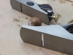 Close-up of two metal hand planes on a wooden surface with wood shavings. A round wooden knob and adjustment knobs are visible on one of the planes.,